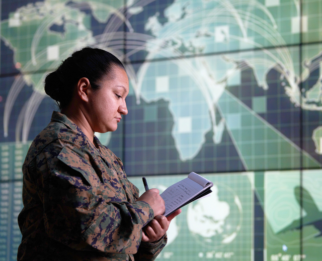 A soldier in uniform writes on a notepad. She is standing in front of a command center with several screens displaying a large global map.