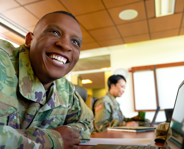 A smiling servicemember in uniform sitting in front of a laptop in a classroom, with another servicemember in the background