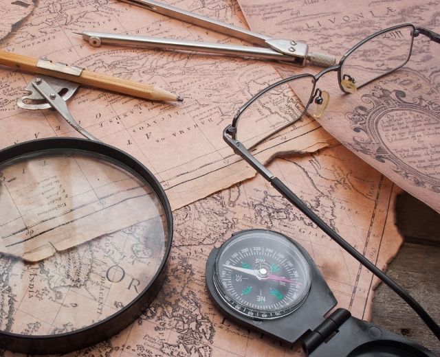A magnifying glass, compass, drafting tools, and eyeglasses rest on vintage maps