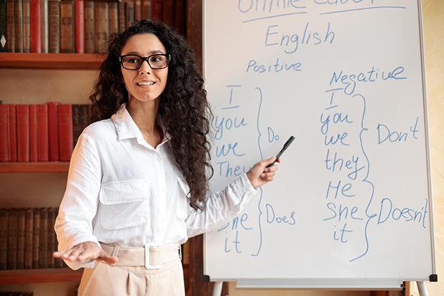 Foreign Language. Portrait of confident smiling young female lecturer wearing glasses teaching English pointing at grammar rules on board with marker, looking back at students at classroom