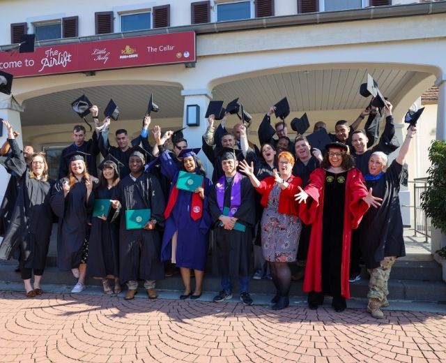 Graduates dressed in caps and gowns celebrate by cheering and raising their caps during a group photo.