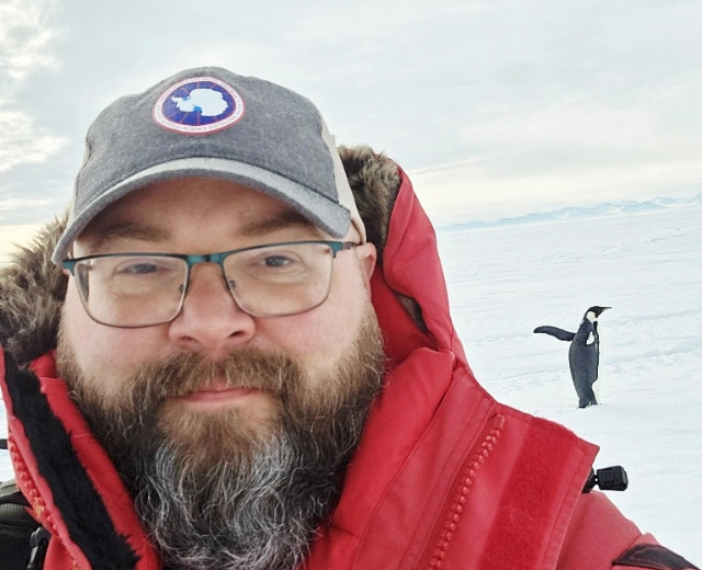 Raymond Polasky outside in Antarctica wearing a hat with the continent on it. A penguin is behind him.