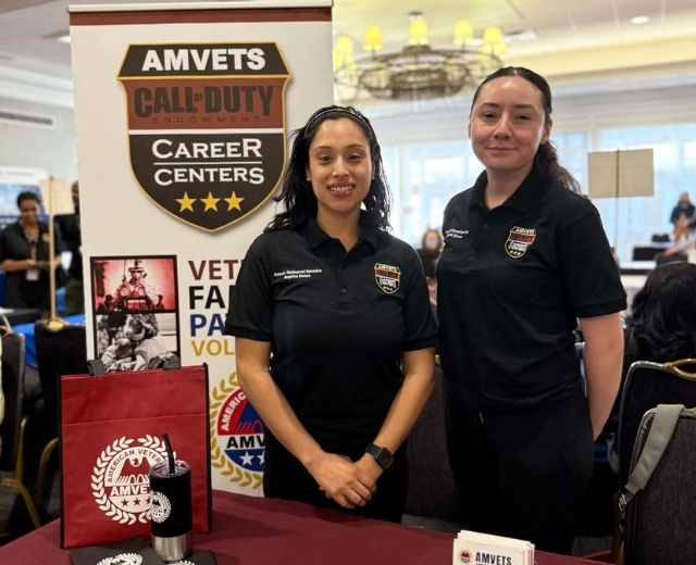 Two women at a booth at a career fair for veterans in front of a sign that reads, “AMVETS Call of Duty Career centers”.