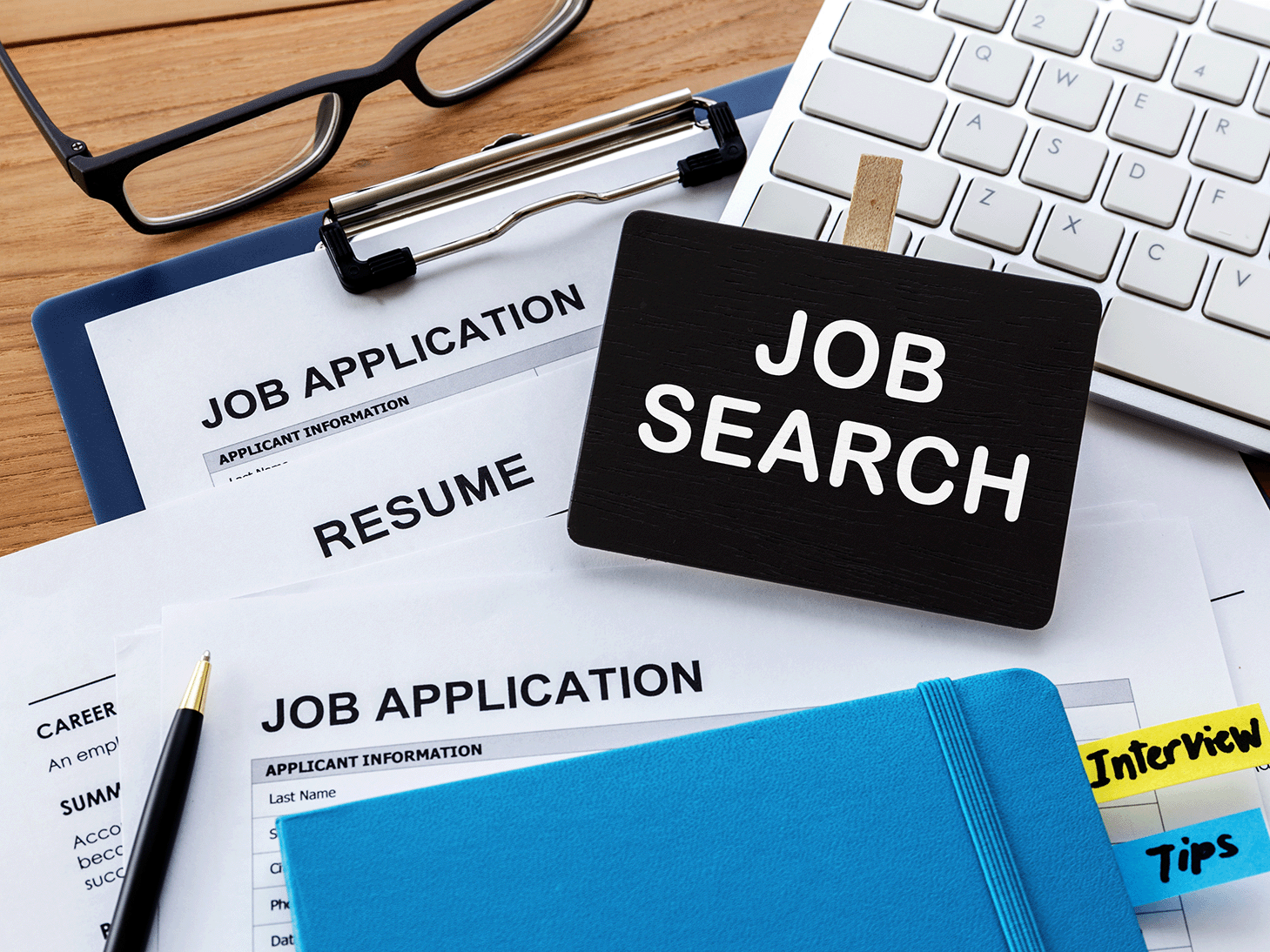 Job applications, resumes, a notebook with 'Interview Tips' and glasses stacked next to a keyboard and tag with the words 'Job Search' on it