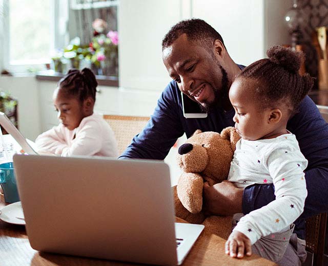 A person on the phone while typing on a laptop and holding their child in their other arm, while another child is looking at a laptop screen in the background.