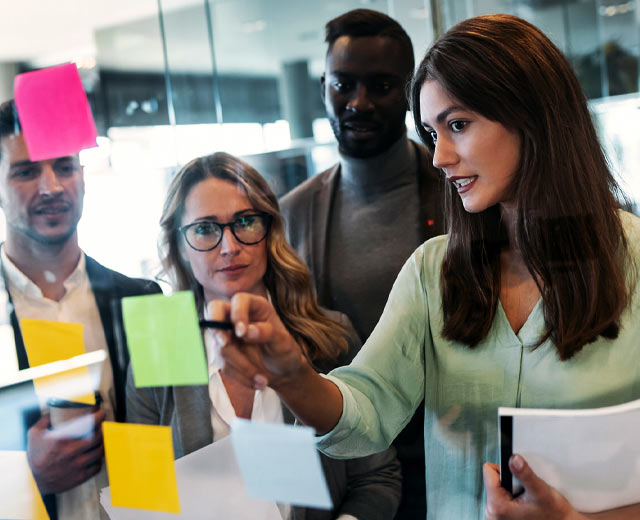 Four people looking at post-its on a clear wall while one of them points with a pen.