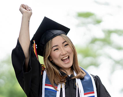 A woman in a graduation gown joyfully raises her arms in celebration of her achievement.