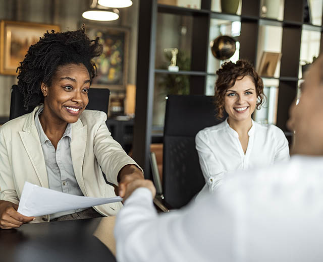 Three people at a desk; two are shaking hands.