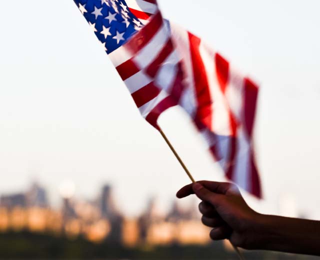 A person's hand holding an American flag with a city in the distance.