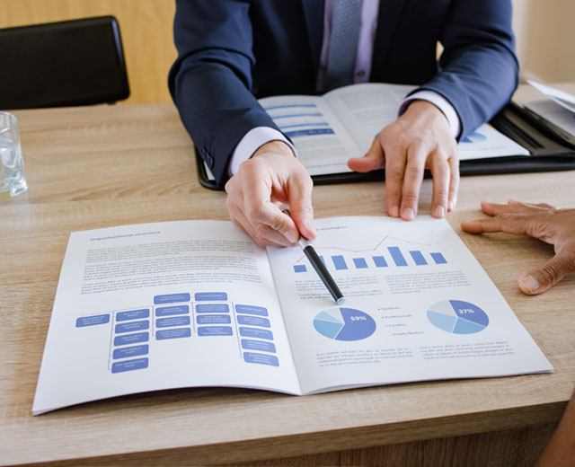 A man is sitting with an open booklet and pointing at a pie chart with a pen.