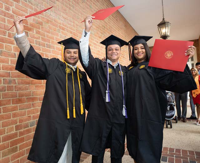 Three graduates stand together smiling and hold their diplomas.