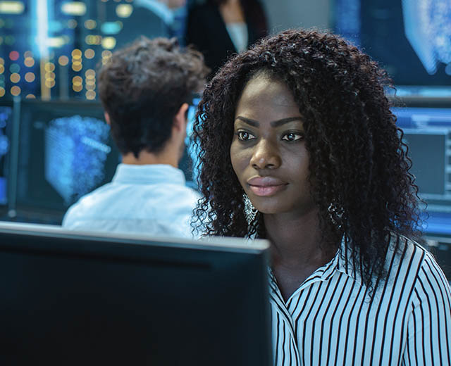 A person working on a computer with multiple people and other computer monitors behind them.