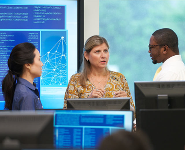 A woman is speaking with another woman and man in a room where there are computers with different graphs.