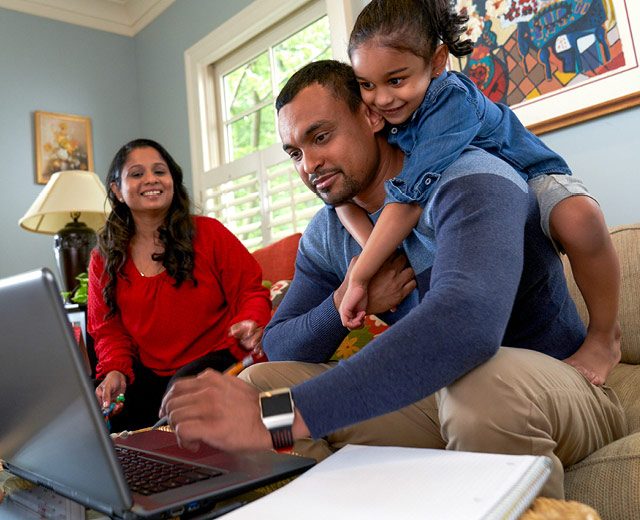 Two adults and a child smiling on a couch while one adult looks at a laptop.