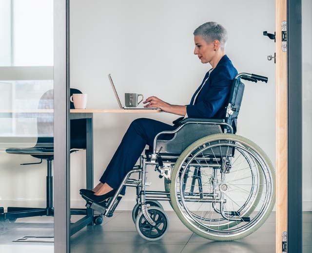 A person in a wheelchair sitting at a table and typing on a laptop.