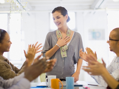 Full image of a woman standing at a table acknowledging the applause of people seated at the table.