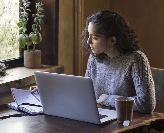 A woman looks at a book next to her while a laptop is open in front of her.