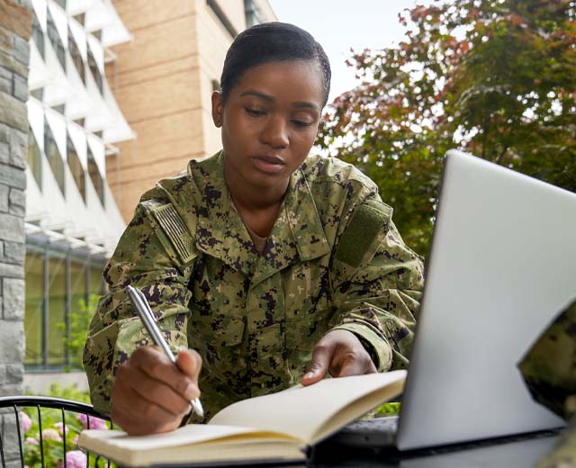A woman in military uniform sits outside, writing in a notebook and looking at a laptop.
