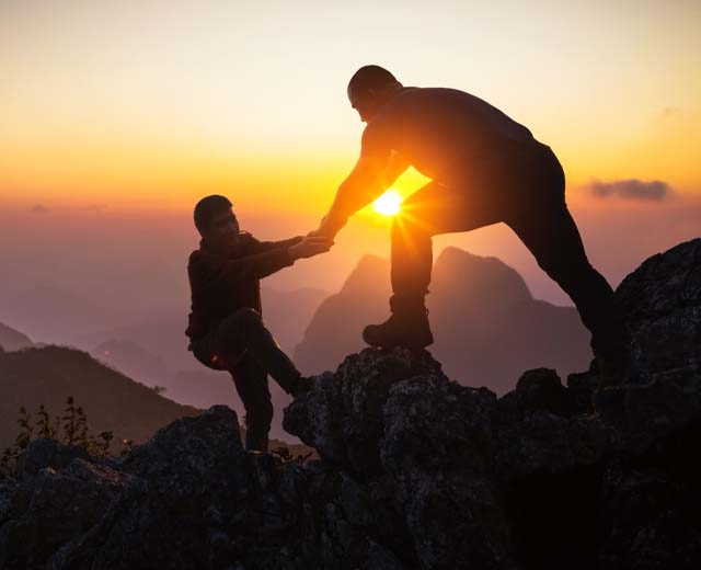 One person helping another climb up the peak of a mountain at sunrise.