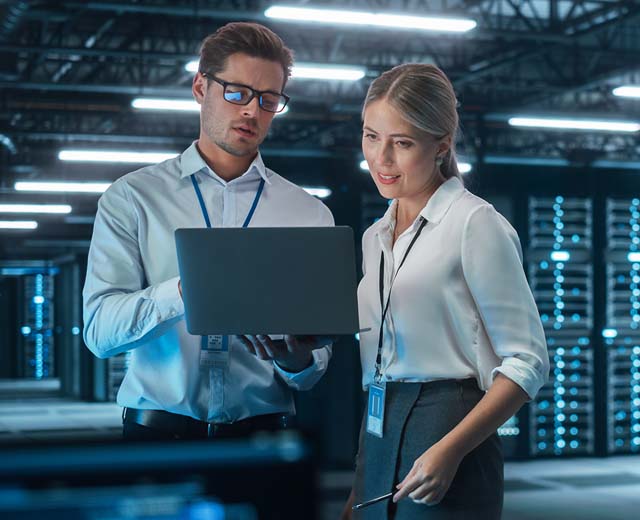 A man and woman look at a laptop together in a building that has mainframe computers in the background.