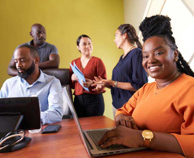 A group of people gather together collaborating in a meeting room. Two are on laptops while three are in conversation. One woman on a laptop is smiling at the camera.