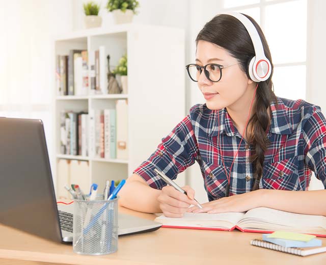 A person with headphones on sitting at a desk looking at a laptop and holding a pen.