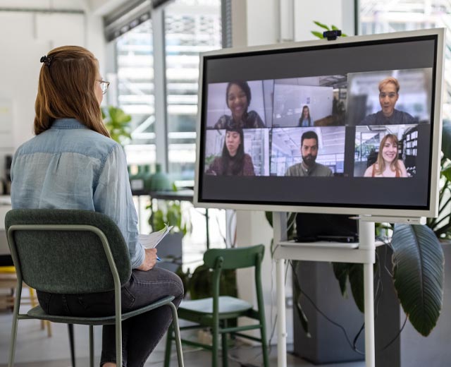 A young woman in front of a large computer screen viewing the faces of six attendees of a virtual meeting.