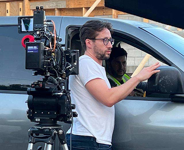 Image of a cameraman standing beside a person in a vehicle. A movie camera faces inside the vehicle.