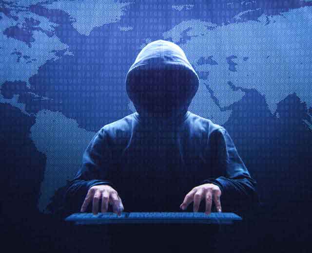 A shadowy figure in a hood typing on a keyboard in front of a map of the world.