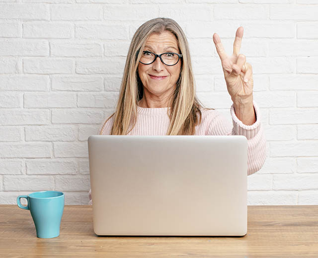 A person sitting behind a laptop screen smiling and giving a peace sign.