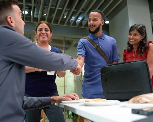 Office colleagues greet each other with a handshake while two coworkers smile nearby.