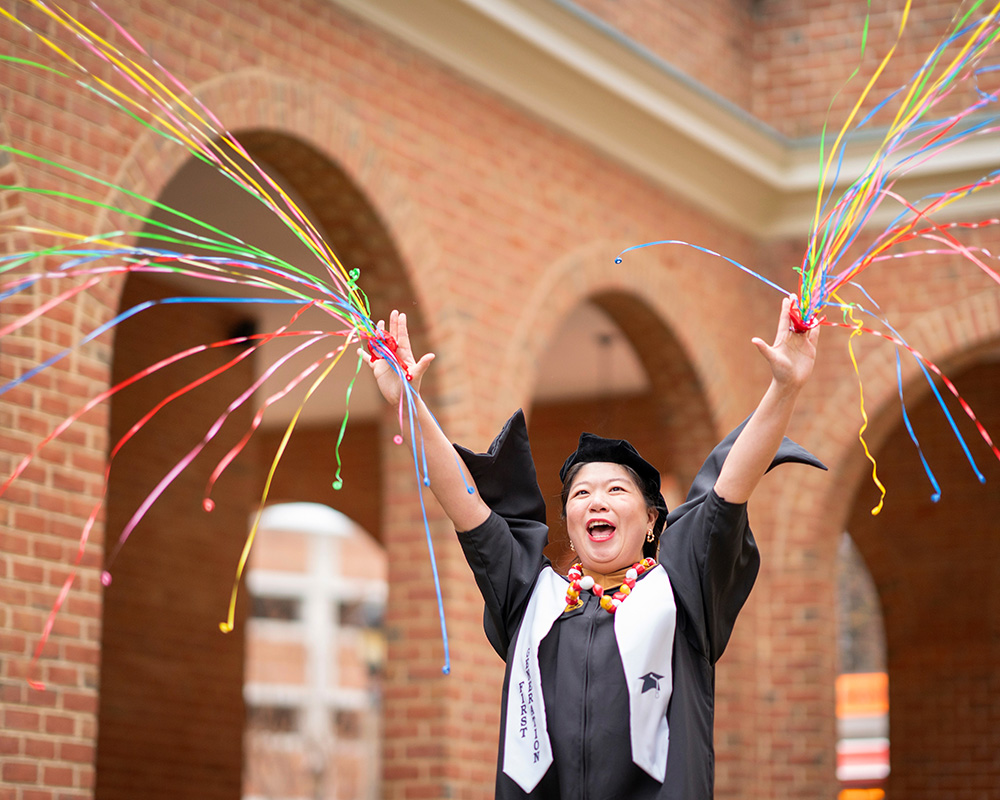 A woman in a graduation gown joyfully holds a vibrant bunch of colored streamers, celebrating her achievement.