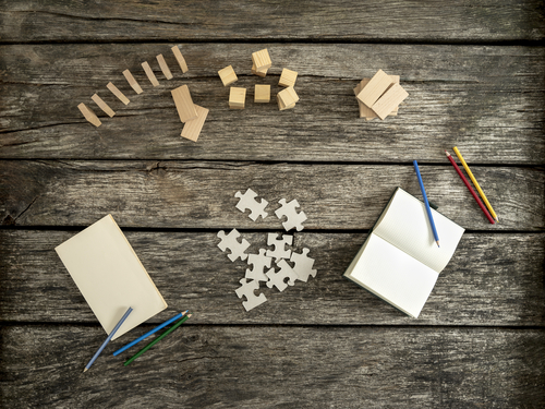 Top view of textured wooden desk prepared for work and exploration - wooden pegs, domino, cubes and puzzles with blank notepads,  paper and colourful pencils lying on it.