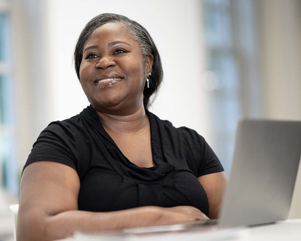 A UMGC graduate in professional clothing smiling and sitting at a desk with a laptop.