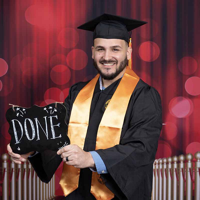 A UMGC graduate in graduation regalia holding a sign that says, "Done."