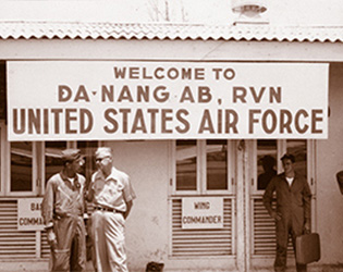 A group of people standing outside a homemade banner that reads "Welcome to Da Nang AB, RVN United States Air Force.