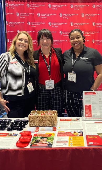 A group of five individuals standing together, smiling in front of a red backdrop with University of Maryland Global Campus logos. They are at a booth displaying brochures, a basket, and promotional items like sunglasses and red socks. They are all are wearing name tags and appear to be representing the organization.