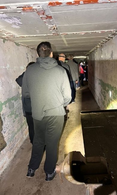 Dr. Michael Mulvey presenting to a tour group outside the historic Kindsbach bunker, which is partially covered in vines, blending in with the surrounding forest. 