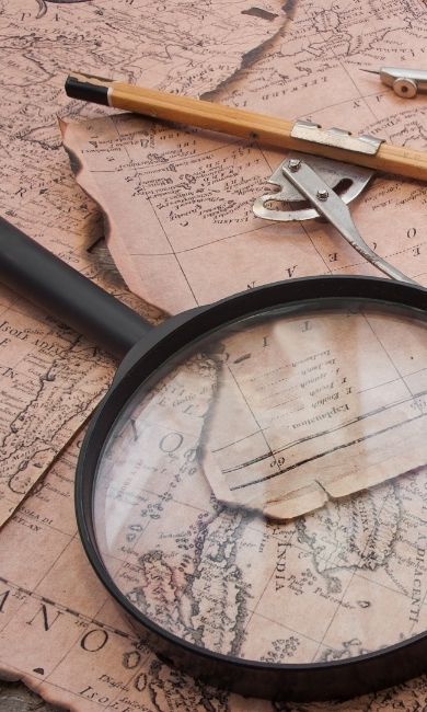 A magnifying glass, compass, drafting tools, and eyeglasses rest on vintage maps