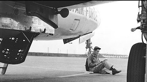 Servicemember leaning against landing gear of airplane studying.