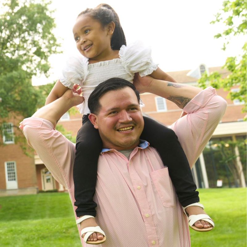 A person outside smiling with a child on their shoulders.