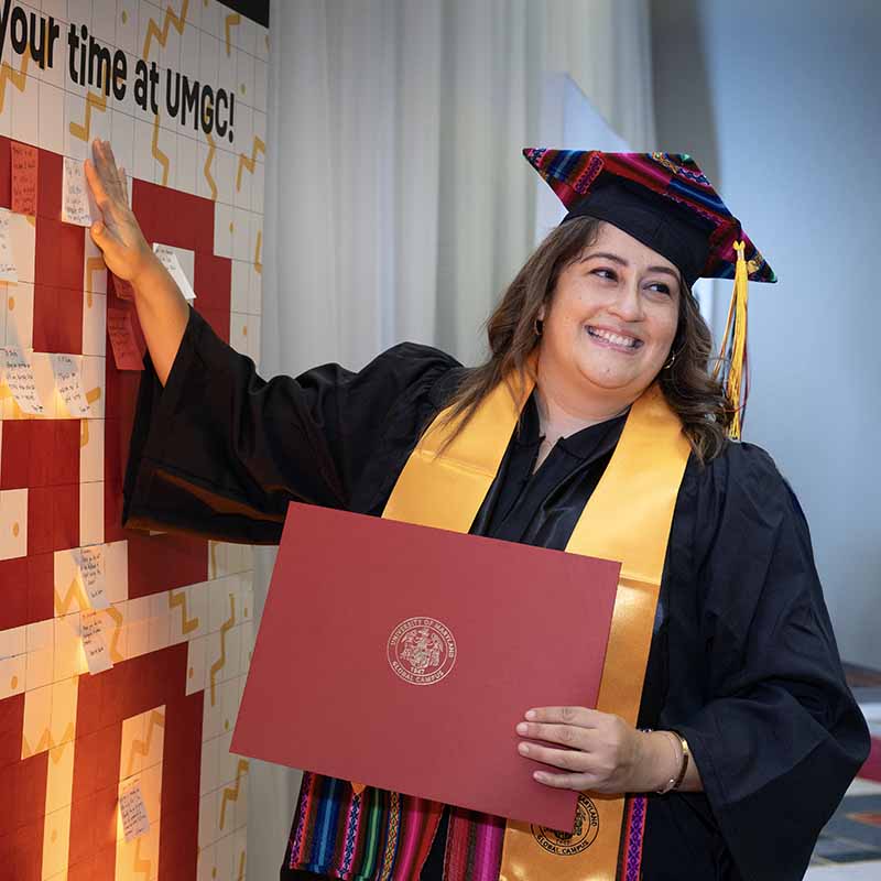 A UMGC graduate in graduation regalia smiling and holding a diploma.