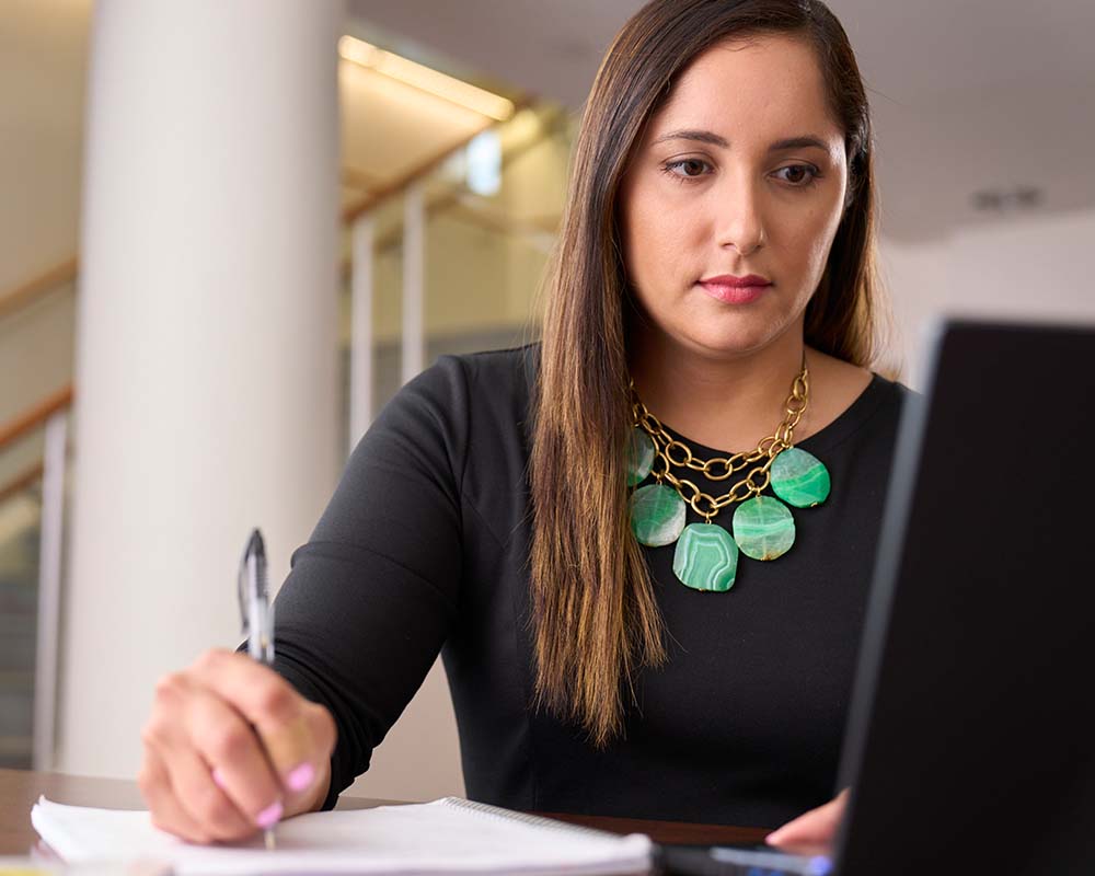 A person taking notes while looking at a laptop screen.