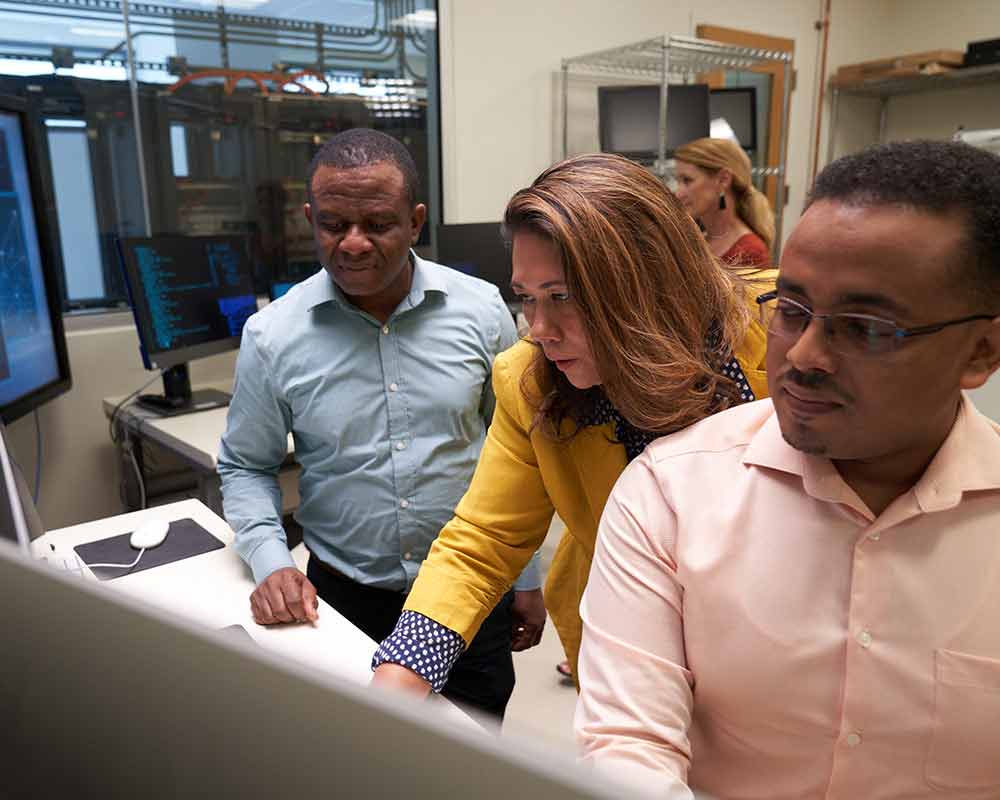 A UMGC student, who is pursuing a master’s degree in information assurance and information security, and another UMGC student, who is pursuing a master’s degree in management and information systems, are looking on as a third person is explaining something. 