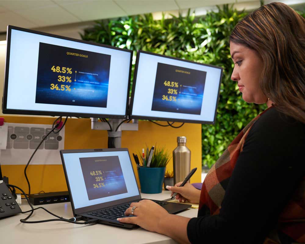 A UMGC student, who is pursuing a bachelor’s degree in psychology, is typing something into a laptop while two screens above them are displaying financial figures. 