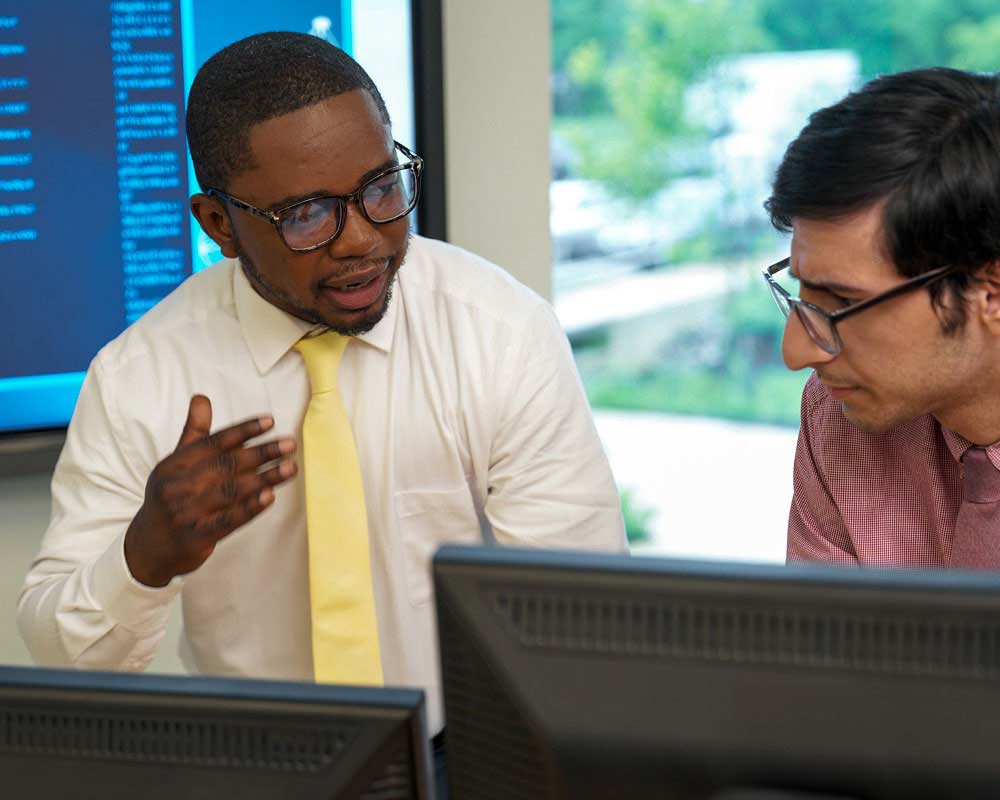 Two UMGC students, one of whom is pursuing both a bachelor’s and master’s in cybersecurity and computer networking and the other of whom is pursuing a bachelor’s degree in cybersecurity and computer networking, are talking while in front of a computer. 
