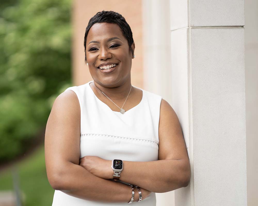 A UMGC graduate in professional clothing smiling and crossing their arms in an outdoor location.