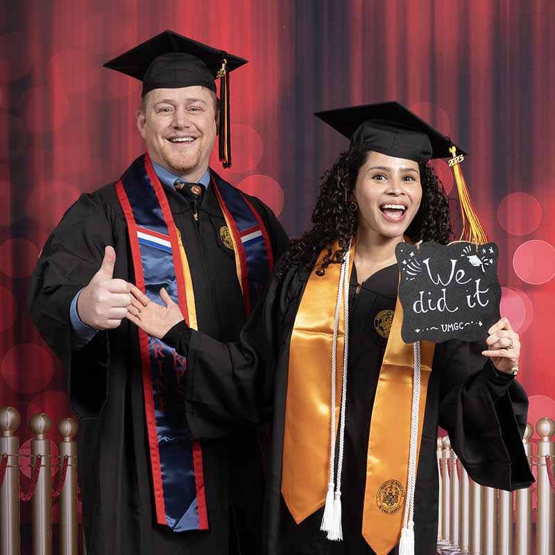 Two UMGC graduates in graduation regalia holding a sign that says, "We did it!"