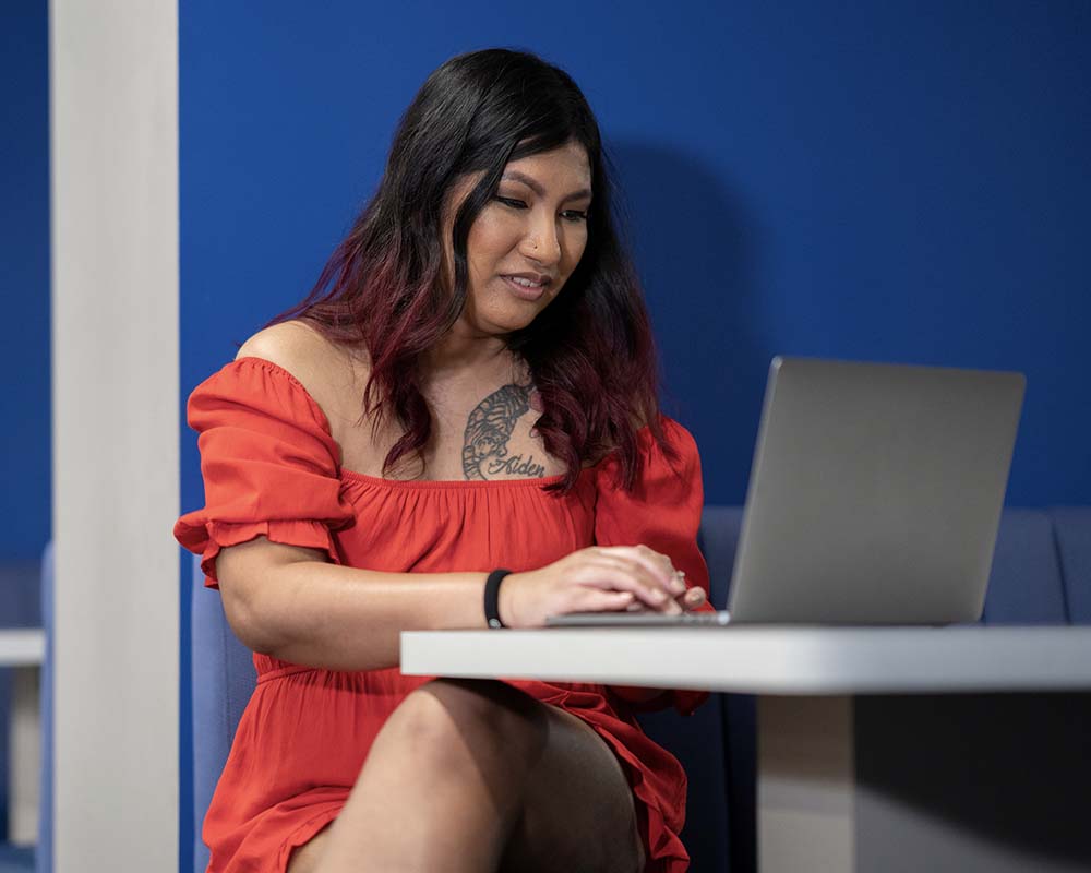 A UMGC graduate in professional clothing smiling and sitting at a table with a laptop.