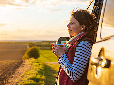A person leaning against a car, holding a mug, and looking out over a country landscape.